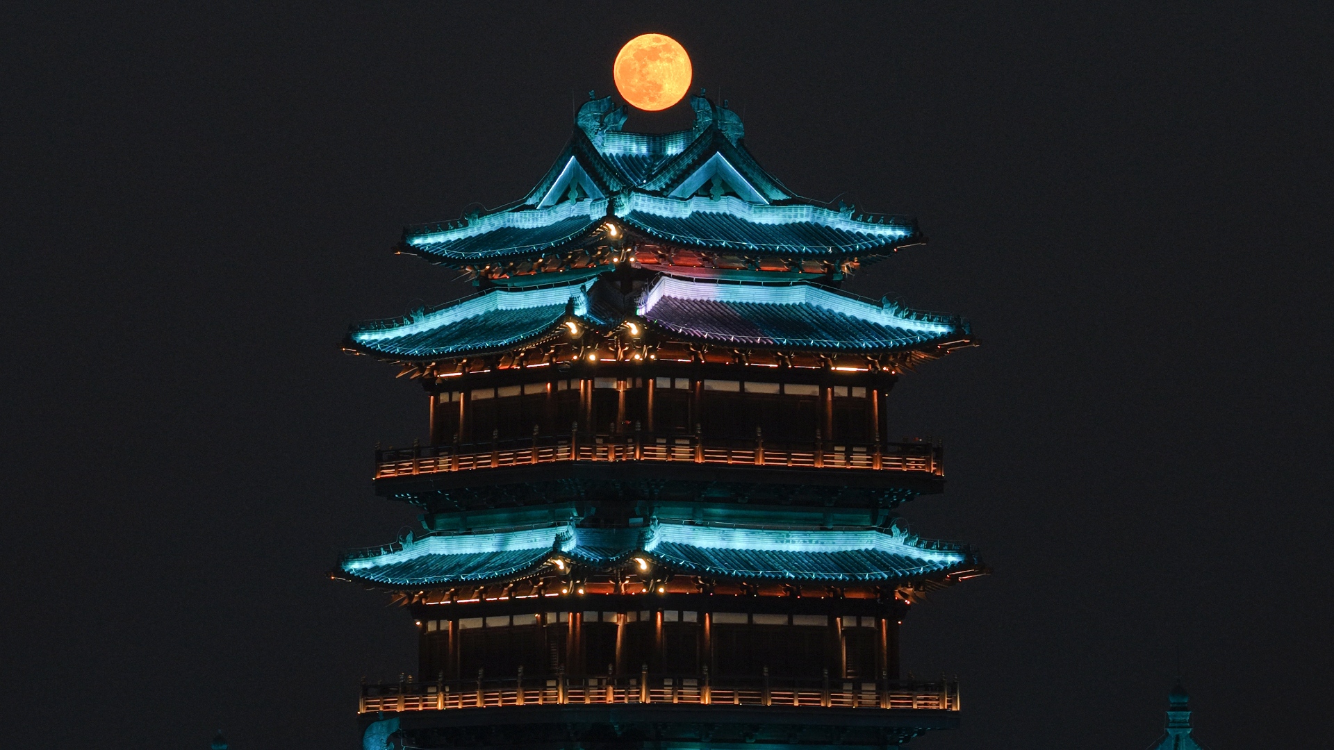 A full moon is pictured glowing above the roof of a traditional Chinese building in a dark night sky. The roof of the building is illuminated to highlight its ornate features.