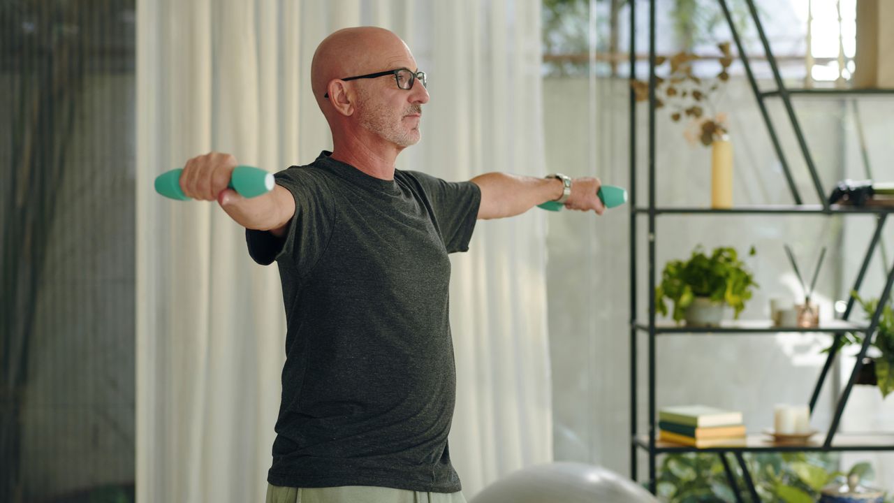 A man performs a dumbbell lateral raise in a living room. He stands with his arms out to the side and light dumbbells in each hand. Behind him we see a curtain and decorative shelving filled with books, plants and candles.