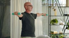 A man performs a dumbbell lateral raise in a living room. He stands with his arms out to the side and light dumbbells in each hand. Behind him we see a curtain and decorative shelving filled with books, plants and candles.