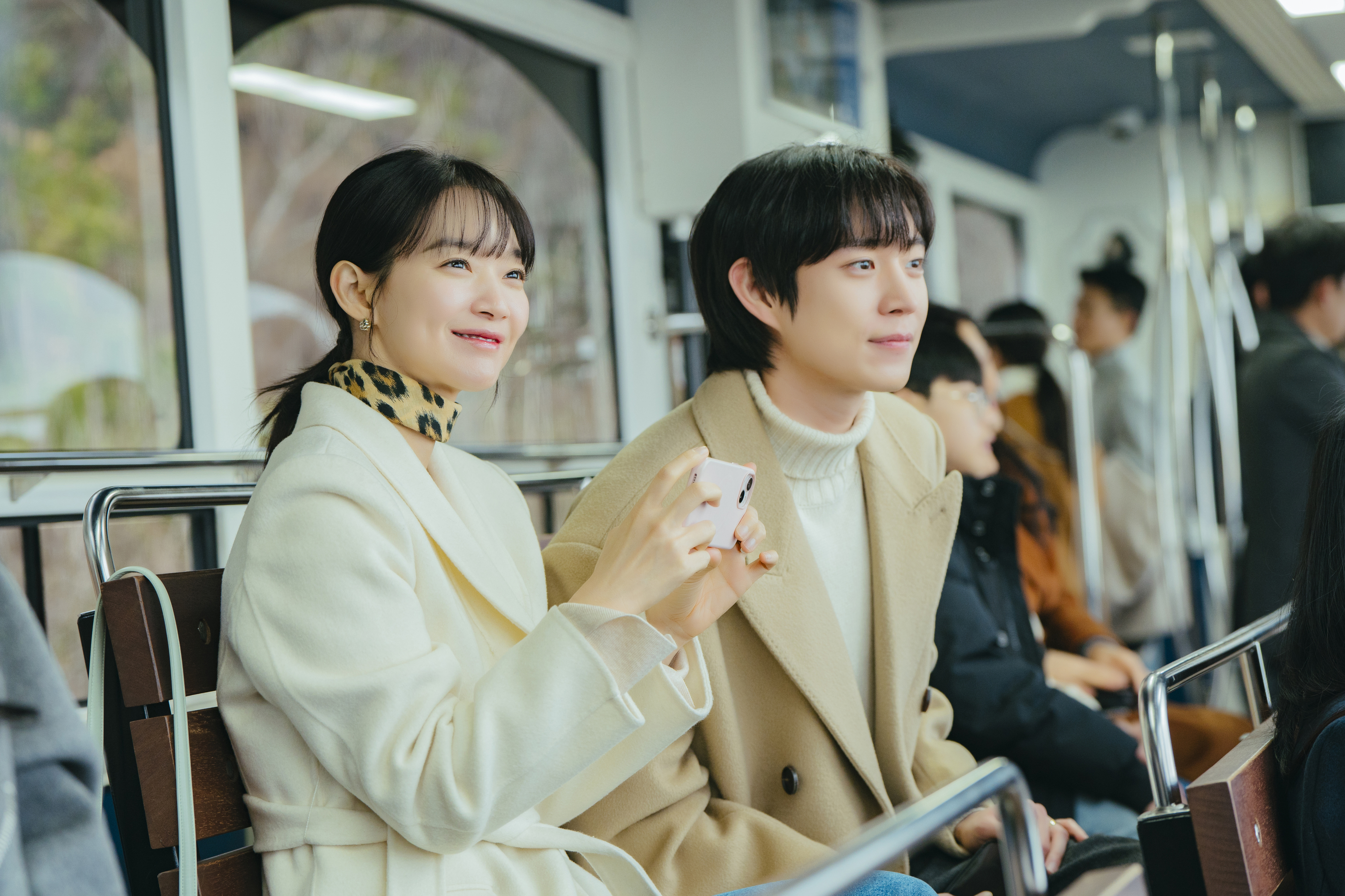 A woman (actress Shin Min-a) holds a phone while sitting next to a man (actor Kim Young-dae). Both wear winter coats while sitting on a bench in a crowded area, in the Prime Video K-drama 'No Gain No Love.'