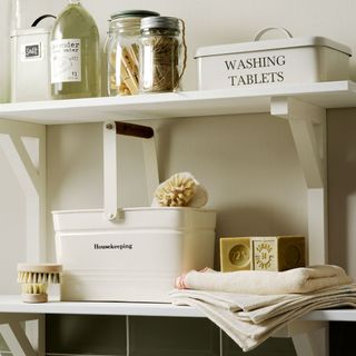 two white shelves in a cream laundry room with an assortment of laundry products including cream housekeeping bucket, washing tablets tin and pegs and twine washing line in jars