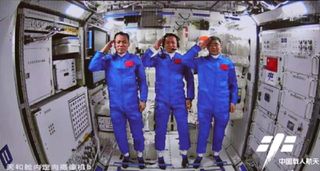Three astronauts wearing blue astronaut jumpsuits salute the camera.