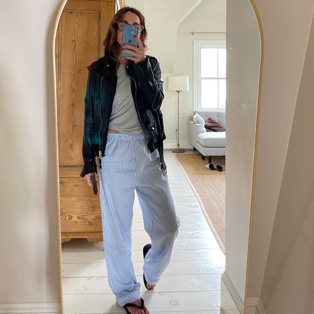 Larissa Mills wearing blue and white striped pajama pants, grey long sleeve shirt, black leather jacket and flip flops in a mirror selfie.