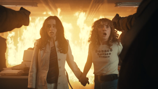 Haley Lu Richardson and Emilia Clarke with guns pointed at them and a fire behind them in Pones