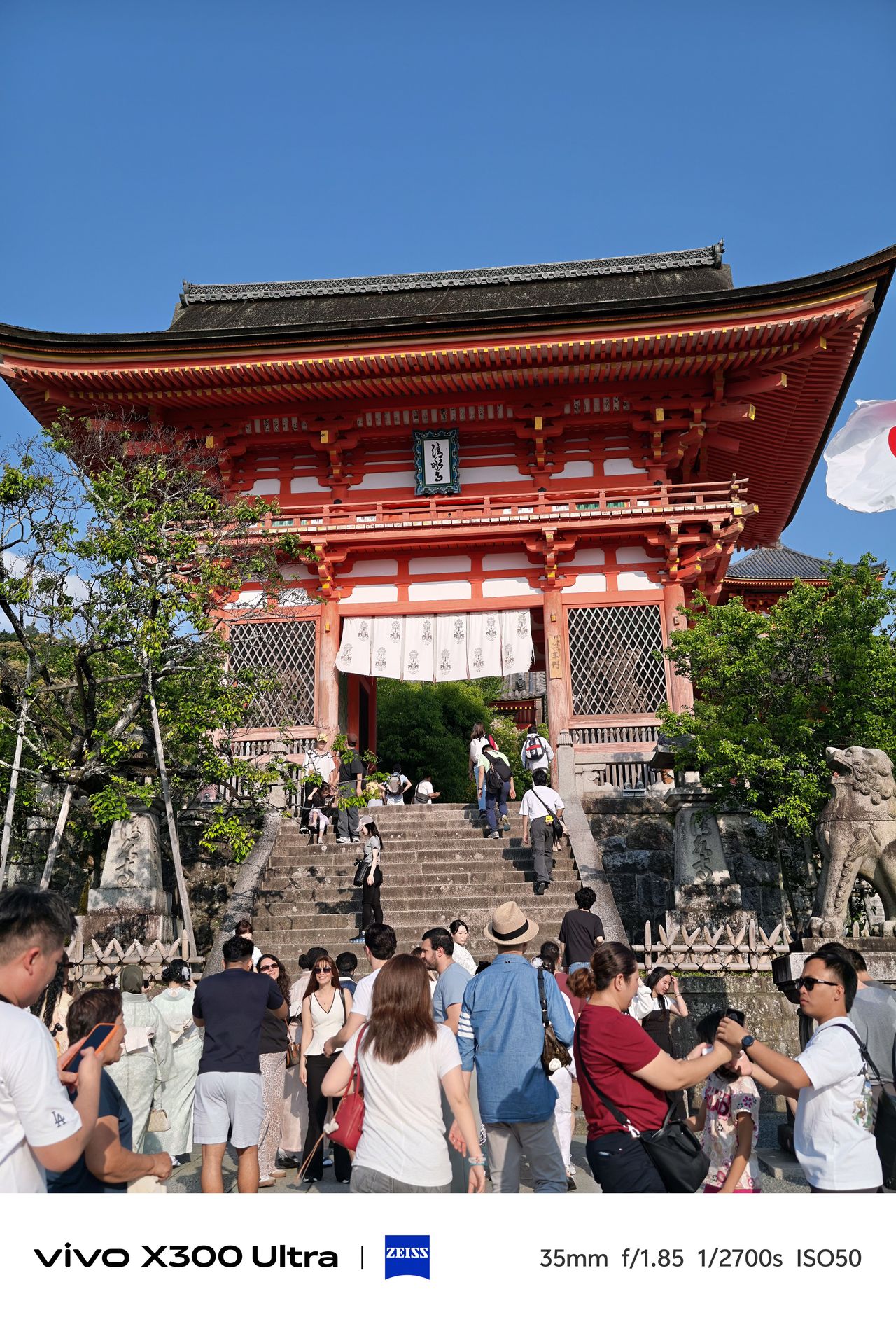 A large crowd gathered at the base of a stone staircase leading up to a grand, multi-tiered orange temple entrance.