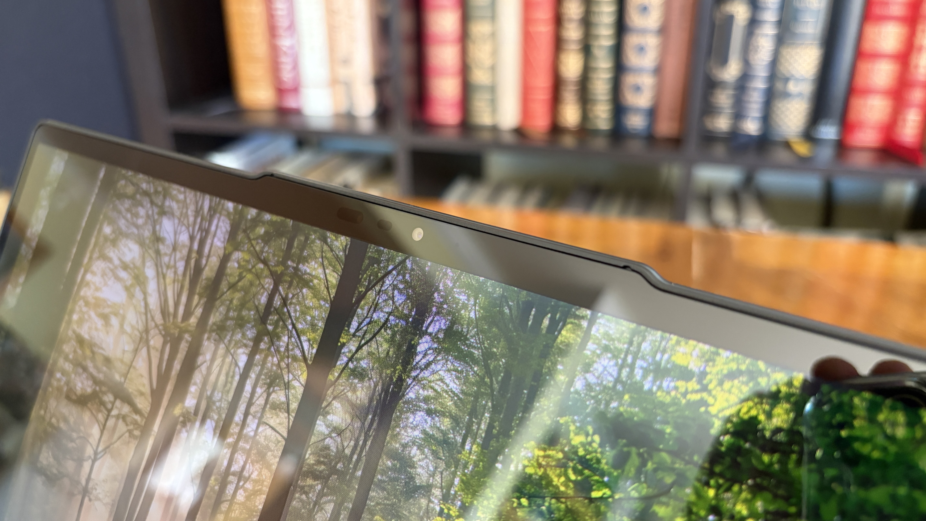 A tablet with a forest scene reflected on its screen is held at an angle. A blurry bookshelf with colorful books is visible in the background.
