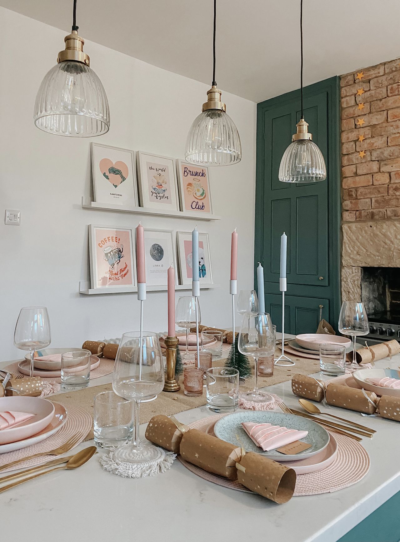 Scandinavian Christmas table setting with pink and blue color scheme, kraft crackers, 3 glass fluted pendants, artwork on a shelf, natural table runner, gold cutlery, pink charger plates