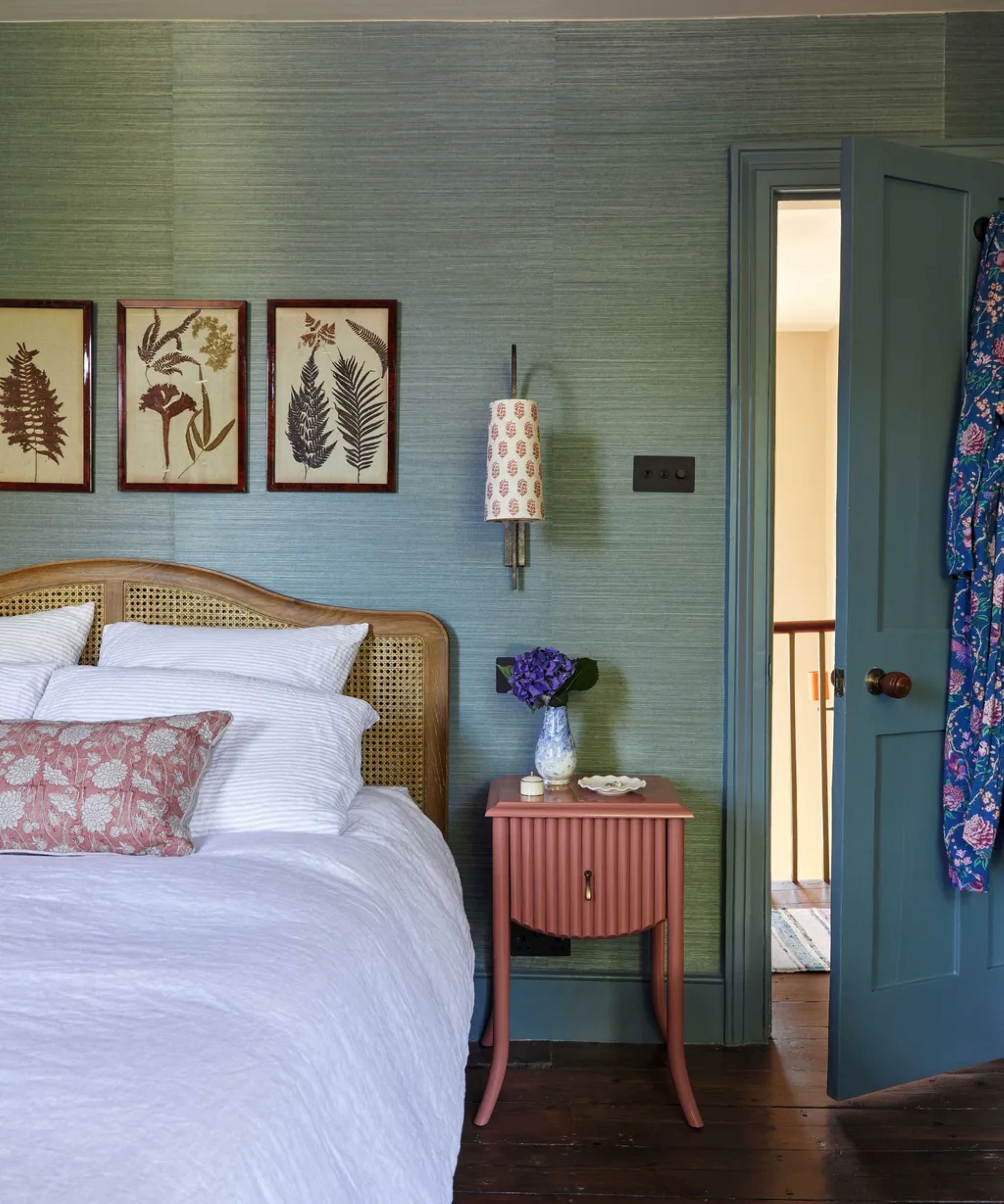 a bedroom with blue grass cloth wallpaper, a pink bedside table, a rattan headboard, white bedsheets, and three pictures hanging above the bed