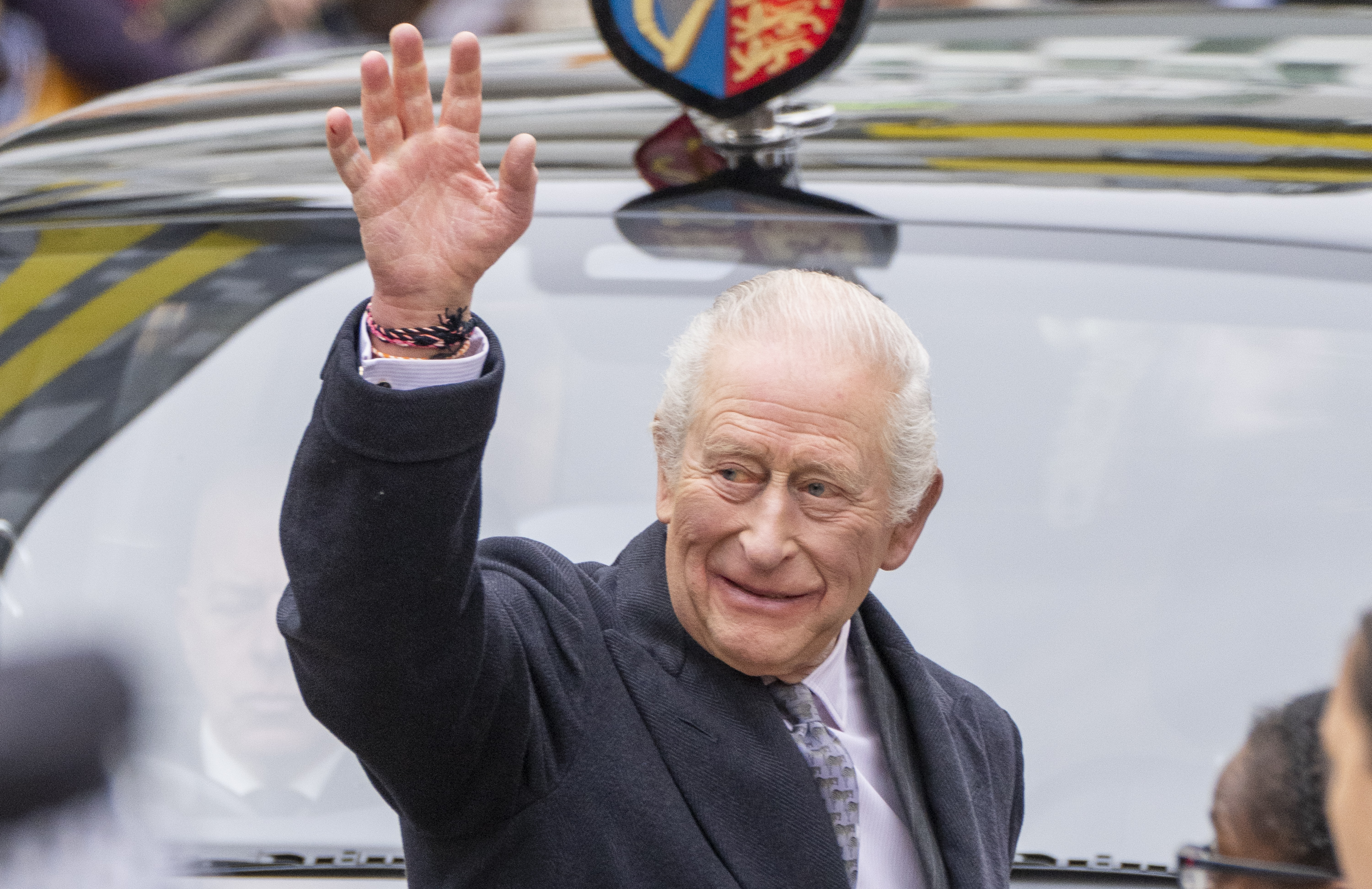 King Charles wearing a gray coat waving in front of a car