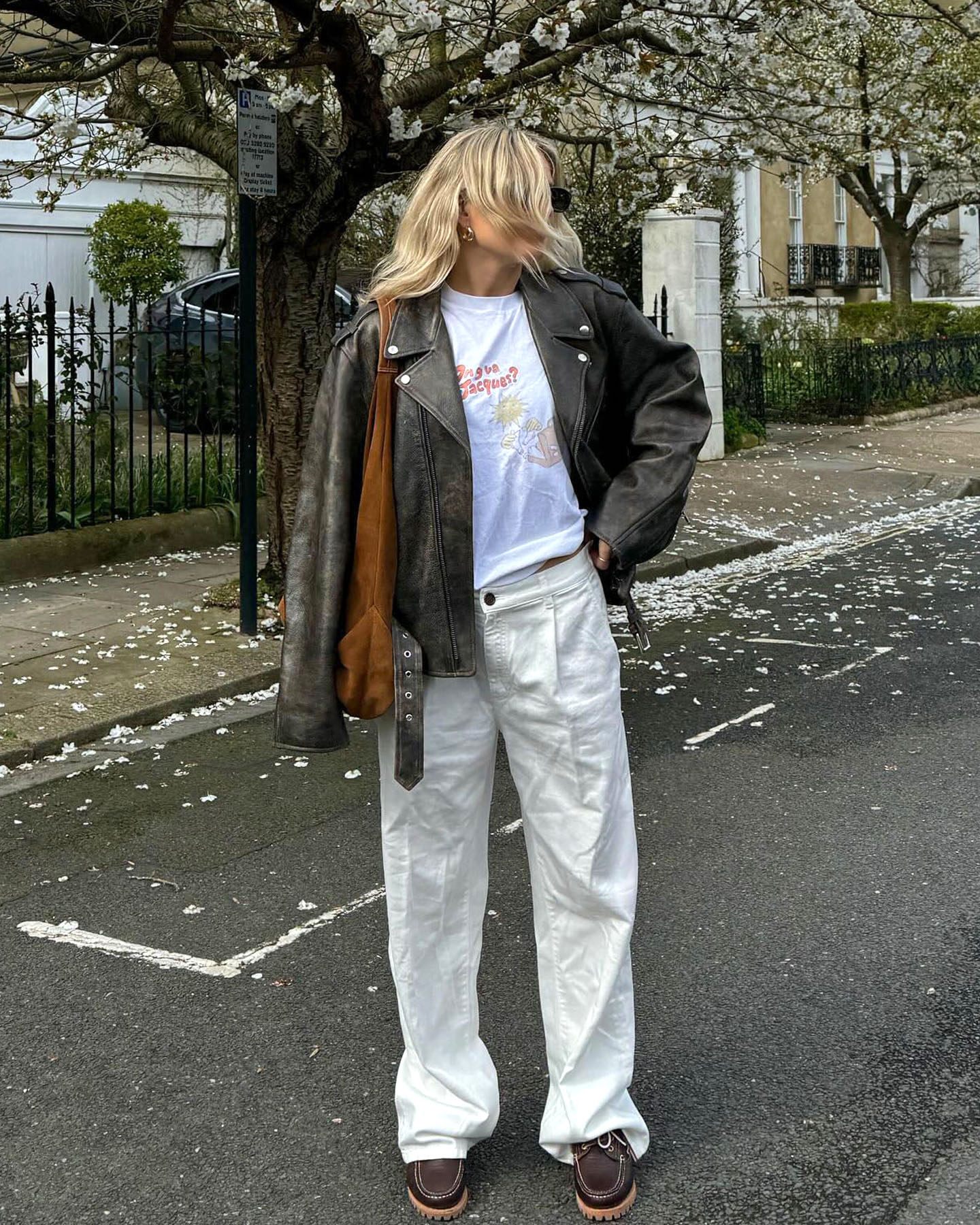 British fashion influencer Lucy Williams wears a stylish outfit with a brown leather moto jacket, a graphic tee, camel suede shoulder bag, neutral baggy pants, and brown boat shoes