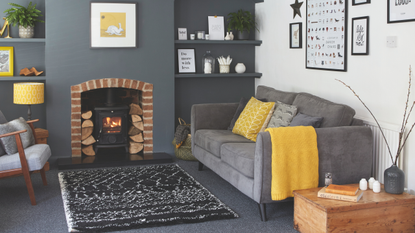 Black and whte rug on a gray carpet. There is a grey sofa with yellow throw and a woodburner fireplace