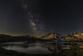 The Milky Way leans to the left of the image center, between peaks of distant mountains, which are reflected in shimmering water in the foreground.