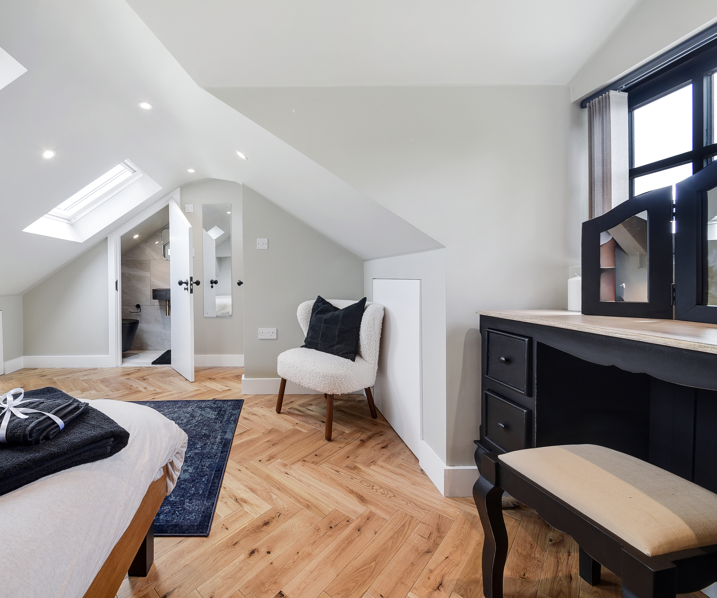 dormer loft conversion bedroom with wooden floor, black dressing table in dormer section, rooflights and door leading into ensuite bathroom