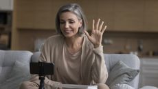 A woman waves as she records a video while sitting on her sofa.