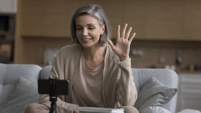 A woman waves as she records a video while sitting on her sofa.