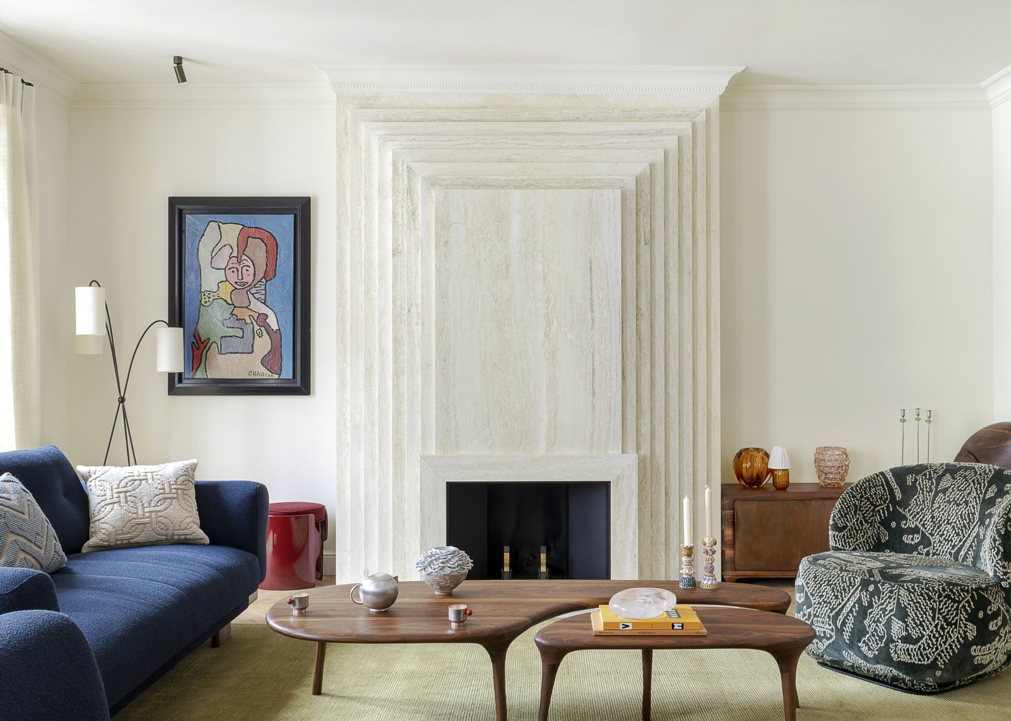 White living room with stone fireplace, pale green rug, a dark blue sofa and dark patterned accent chair, and sculptural wood coffee tables