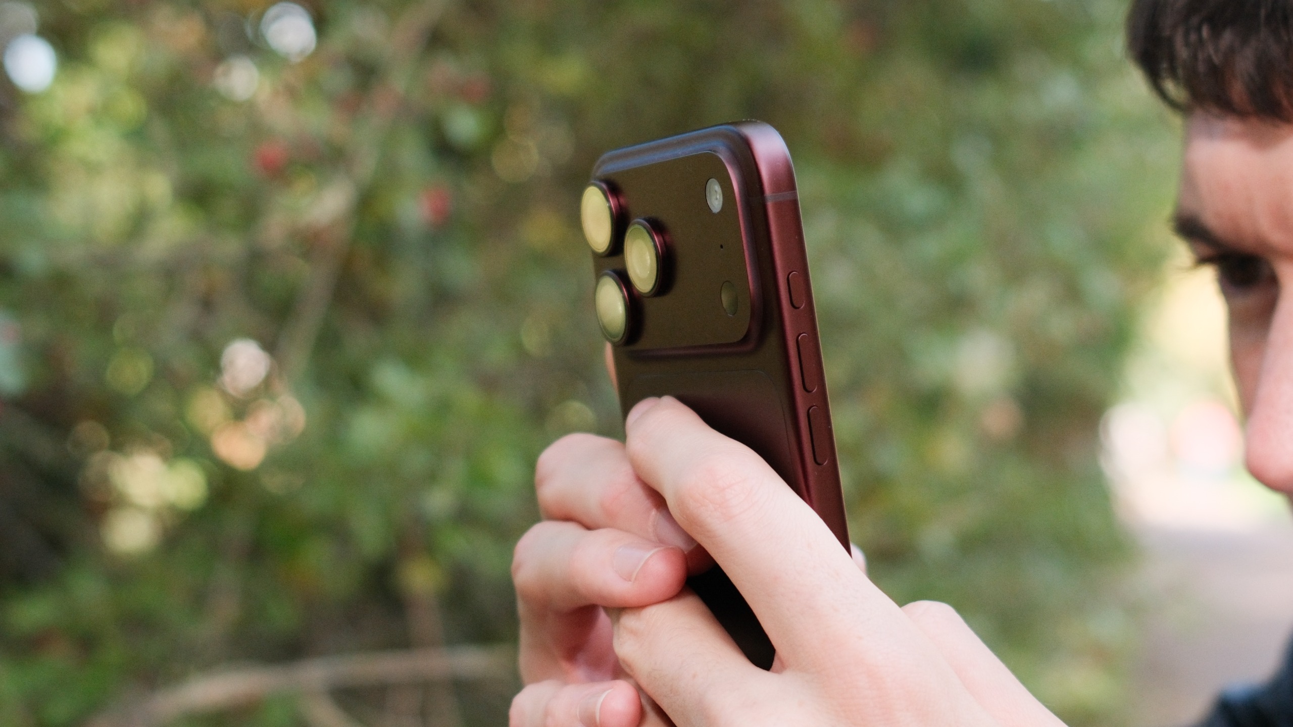 Person holding a burgundy iPhone 18 Pro Max outdoors, with the triple rear cameras facing toward the subject.