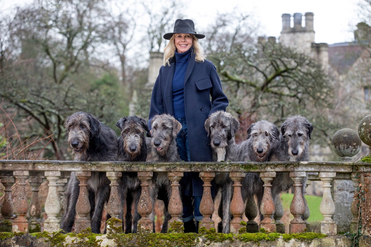 Trudie Styler poses behind a moss-covered stone balustrade with six Irish Wolfhounds lined up in front of her. She wears a navy coat and fedora, with a grand house and wintry trees in the background.