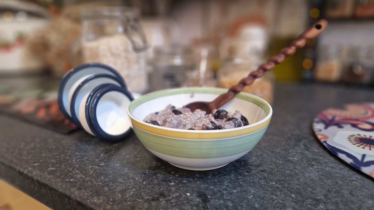 A bowl of oats sit on a kitchen counter in the foreground. Out of focus in the background are ingredients in glass jars and a set of enamel measuring cups.