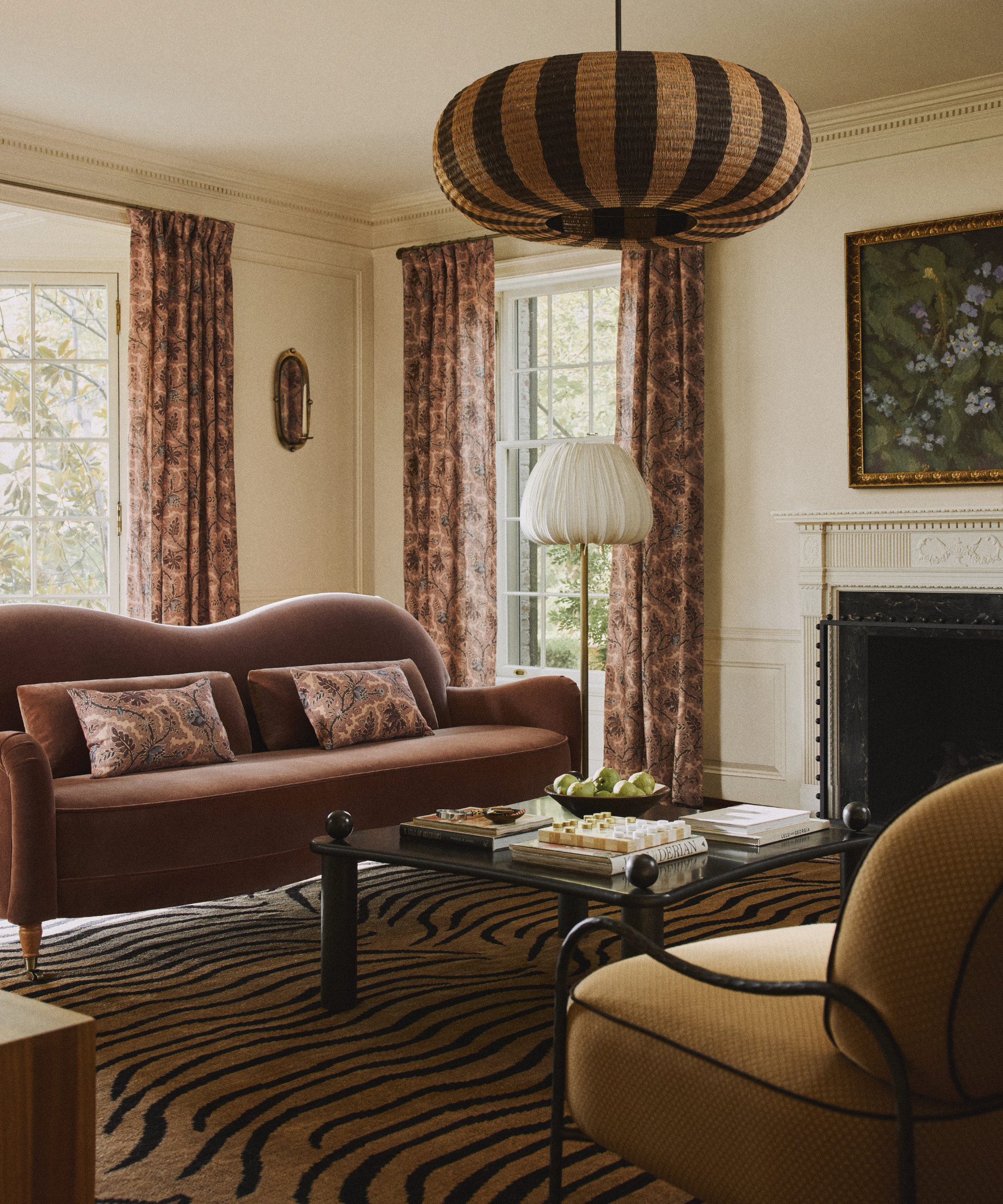 a cozy living room with a tiger striped rug, a mauve couch with pillows on it, large windows with floral printed drapes, and a striped woven pendant light
