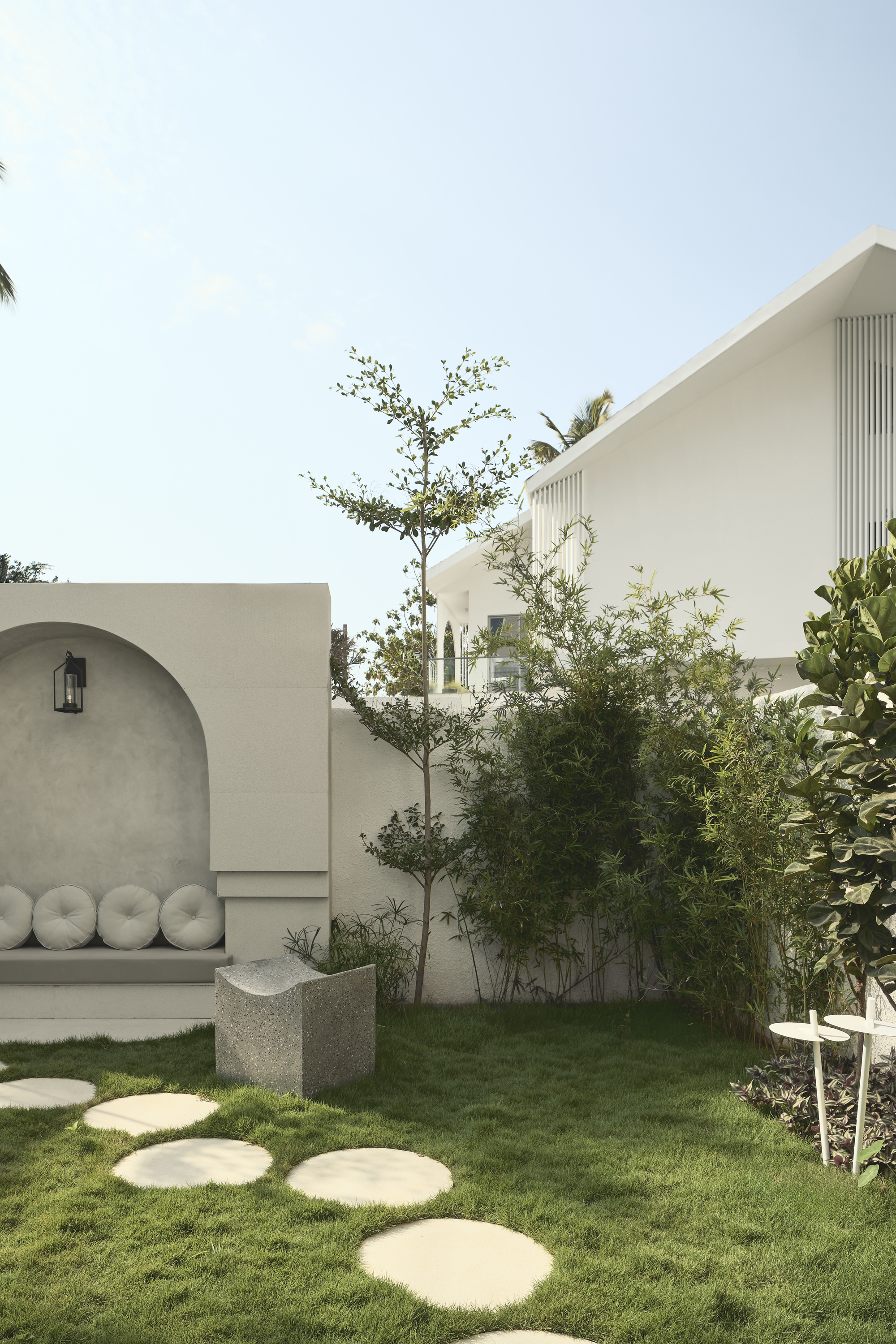 Image of a garden lawn with stepping stones that lead to a modern, gray-beige bench seat built into an exterior wall. There are small trees planted along the wall as well.