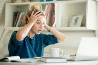 Woman at table with cup and laptop computer grabbing her head, with her eyes wide. Bookshelf in the background. 