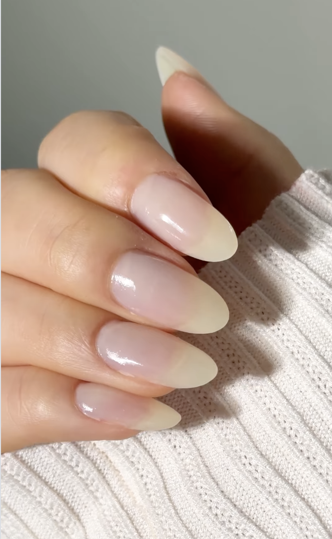 a hand with long almond nails with a barely-there polish and a white sweater