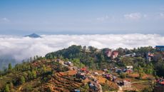 An aerial view of a mountain top village with small colorful houses surrounded by ledges and trees, with clouds in the distance. 