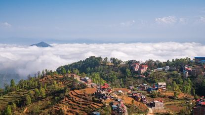 An aerial view of a mountain top village with small colorful houses surrounded by ledges and trees, with clouds in the distance.