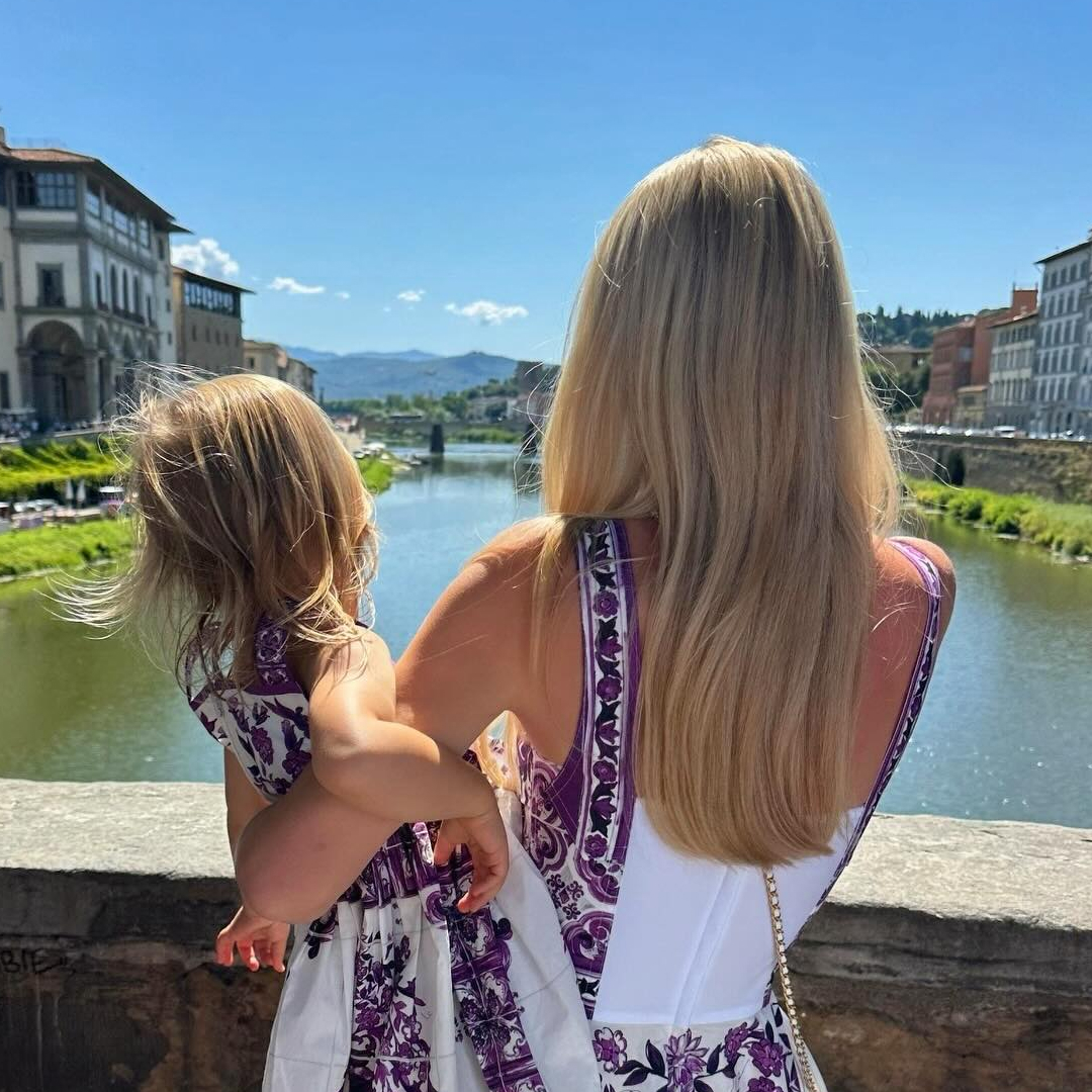 Kitty Spencer holding her daughter Athena on a bridge looking at the water, both wearing white and purple dresses