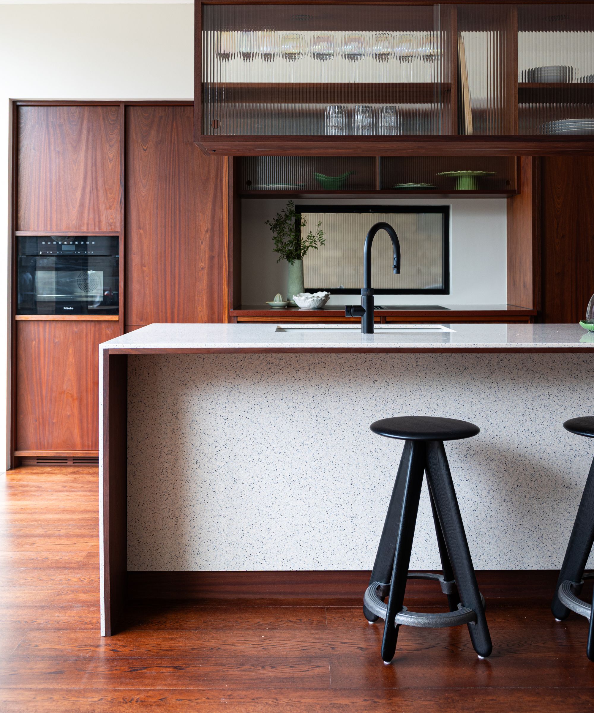 Walnut kitchen island with speckled terrazzo sides, black bar stools, glass-front cabinets above sink.
