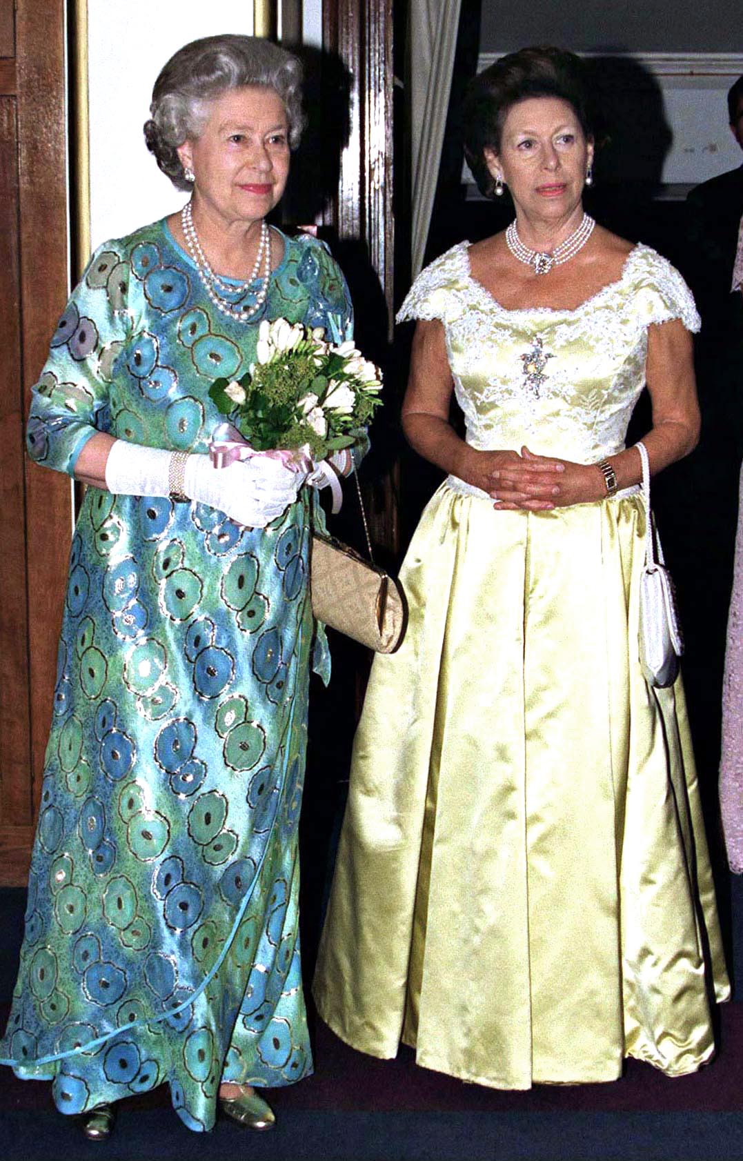 Queen Elizabeth wearing a purple and green gown standing next to Princess Margaret in a yellow gown