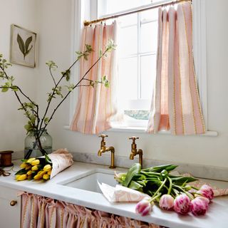 pink cafe style curtains in a white kitchen with brass taps and flowers by the sink.jpg
