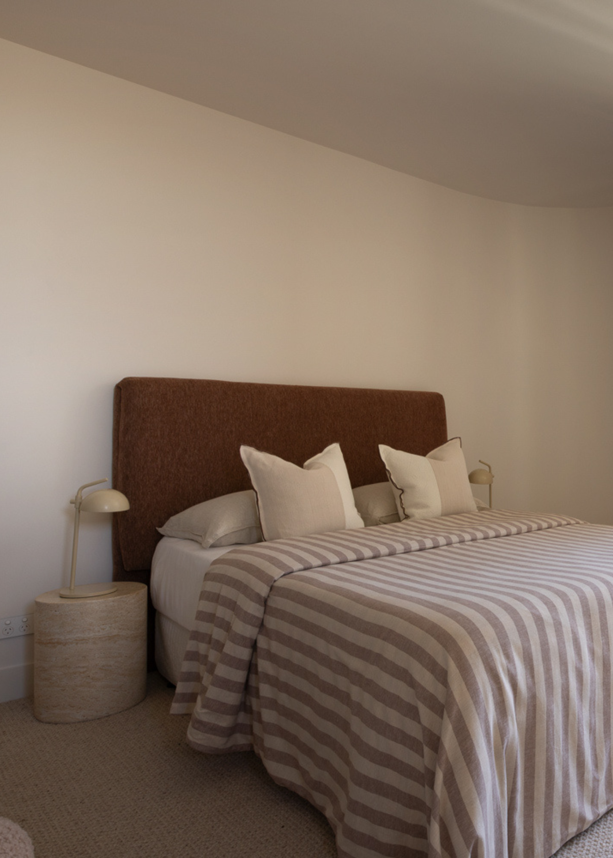 A white bedroom with a stone side table and a table lamp, a brown headboard, a striped bedscape, and color-blocked pillows