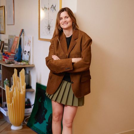 French fashion creative Eugenie Trochu poses in her Paris apartment wearing a brown suede blazer jacket, a black top, and a pleated olive green mini skirt