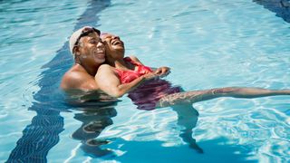 Man and woman in swimsuits, goggles and hats laughing and swimming in the pool