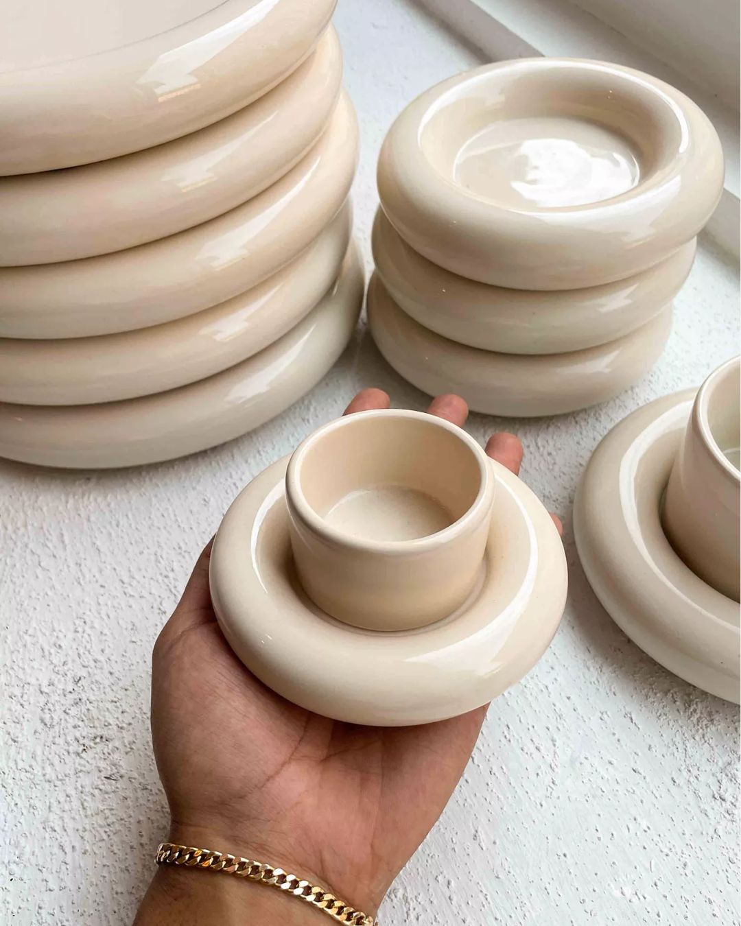 Chunky Earthenware Cup and Saucer in Cream, held by a hand wearing a golden bracelet in front of other similarly colored sauces and cups.
