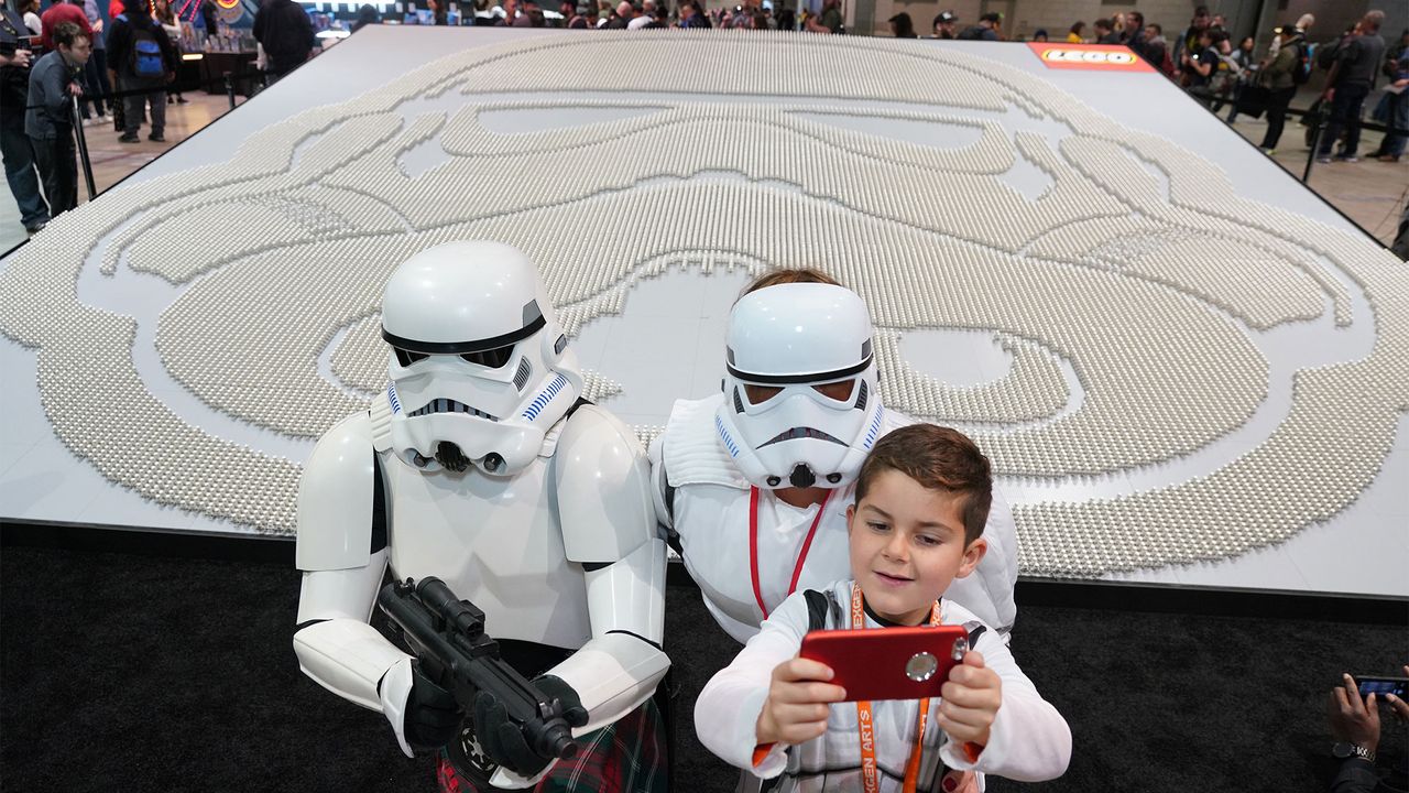 At Star Wars Celebration in Chicago, an attendee takes a selfie with two Stormtroopers in front of the Guinness World Record-breaking display of Stormtrooper minifigures, which stand in a formation representing a Stormtrooper's helmet.