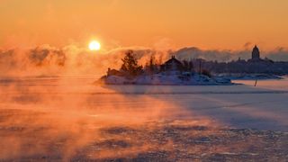 A serene winter landscape featuring a rising sun casting orange hues over a frozen lake, with mist enveloping a distant snowy island and buildings