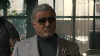 Sylvester Stallone with a goatee, wearing sunglasses and a black turtleneck and gray jacket in Tulsa King. 