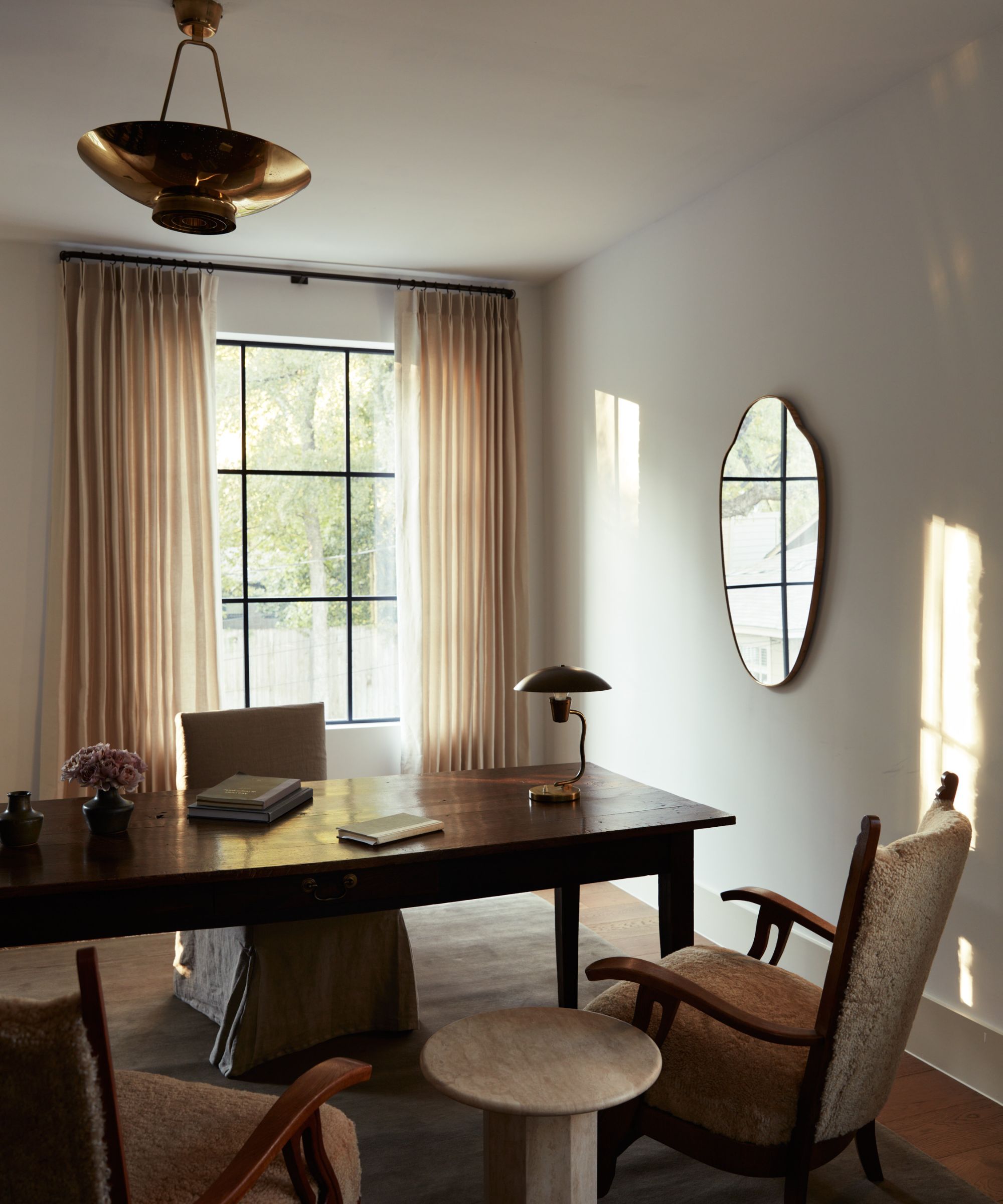 Cozy neutral study with wooden desk, two chairs and side table in front of desk, sheer curtains on window behind, mirror to the right on the wall