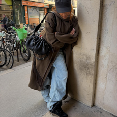 Fashion person @mv.tiangue wears a Gen Z spring outfit idea. She wears a brown wool scarf coat, baggy jeans Beckett wedge trainer and a baker boy hat. This image was taken from her Instagram recently.