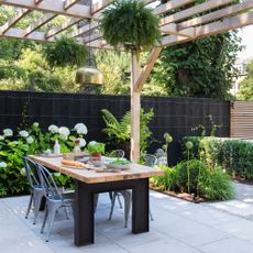 Patio with dining table with black legs and wooden top, metal bistro style chairs and a gold pendant light hung above from a wooden pergola