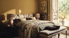 A yellow painted bedroom with a green floral bed. A grey upholstered bench at the base, and a vase of cherry blossom stems in the window.