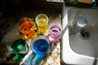 Different glass pots containing separate yellow, green, blue, orange and purple dip dye on a kitchen side next to a sink.