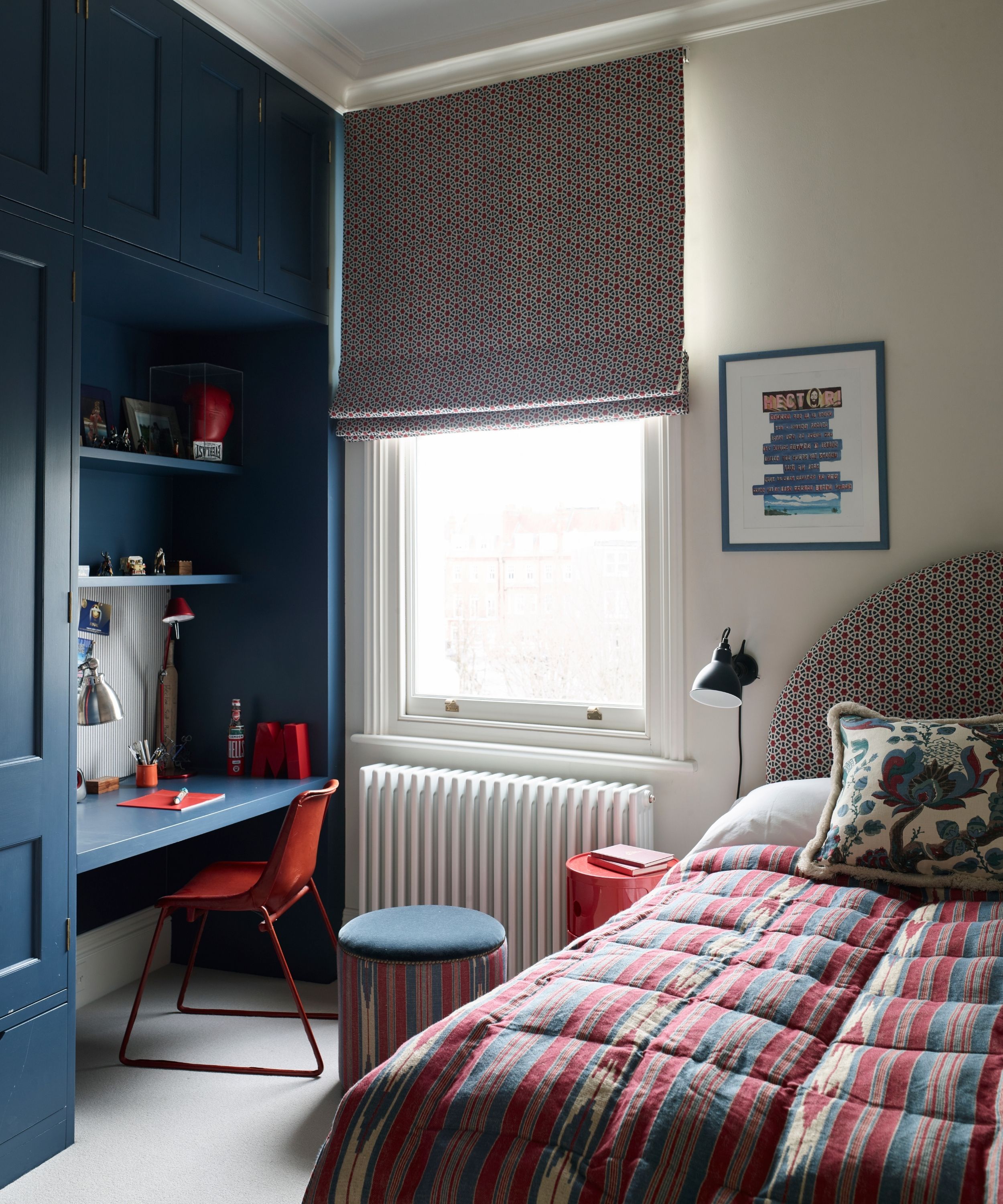 White bedroom with blue desk and red chair, single bed with patterned headboard, cushion and quilt, and large window with a matching patterned blind, white radiator, and small stool.