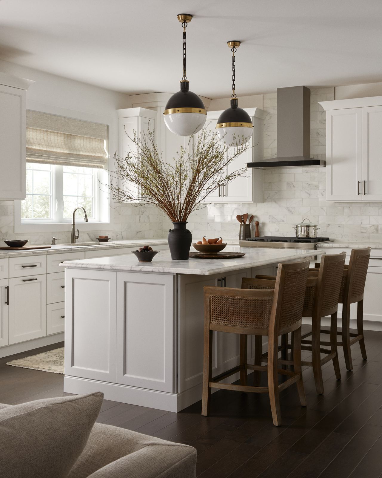 Image of a modern kitchen with true white cabinetry and a matching kitchen island. The countertops are white marble, and the backsplash is white zellige tile. There are dark hardwood floors and brown rattan barstool chairs at the island.