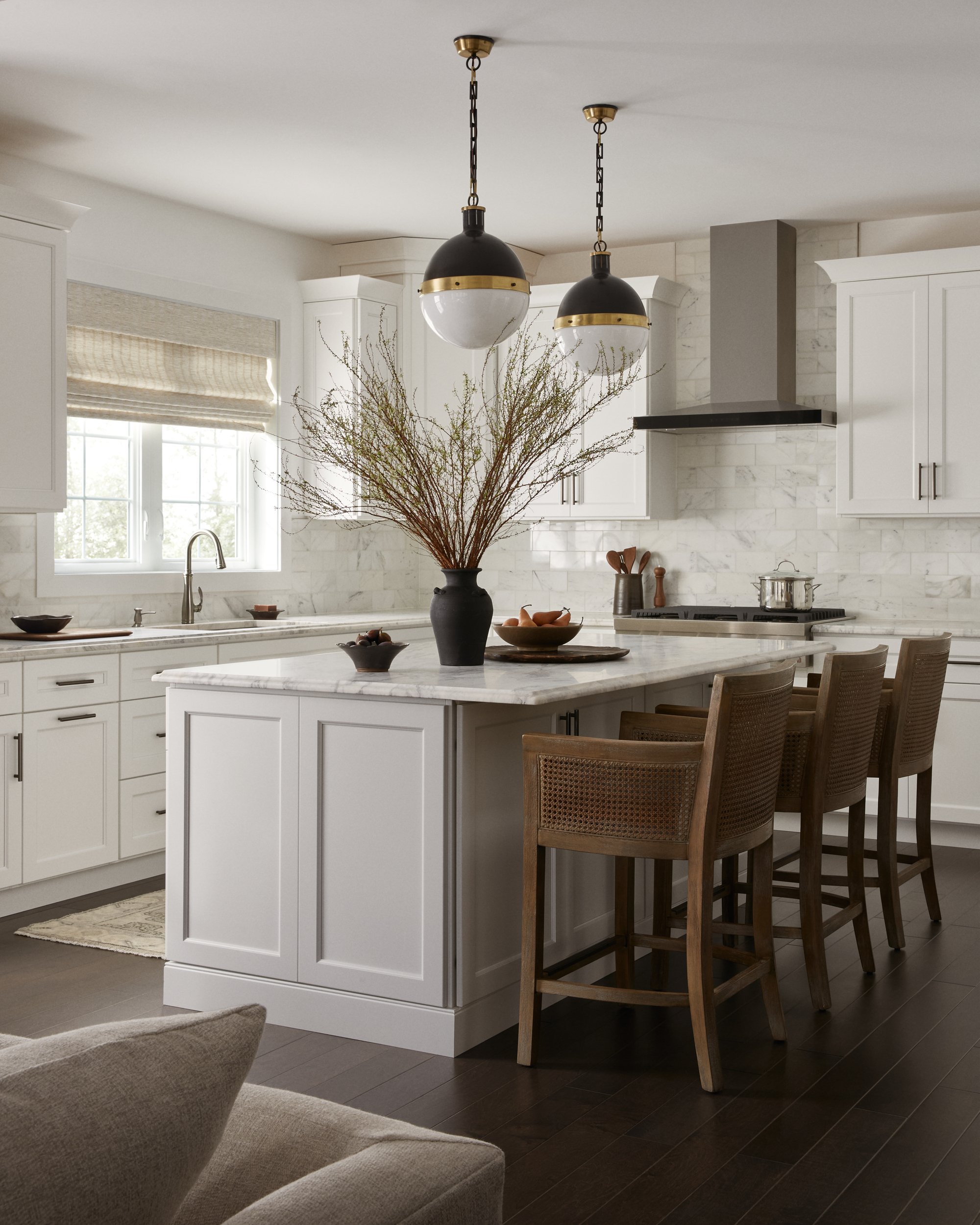 Image of a modern kitchen with true white cabinetry and a matching kitchen island. The countertops are white marble, and the backsplash is white zellige tile. There are dark hardwood floors and brown rattan barstool chairs at the island.