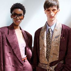 A female and male model stand in a beam of sunlight, the couple are both sharply dressed in relaxed tailoring, their suits are in a matching maroon fabric.