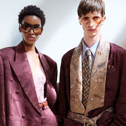 A female and male model stand in a beam of sunlight, the couple are both sharply dressed in relaxed tailoring, their suits are in a matching maroon fabric.